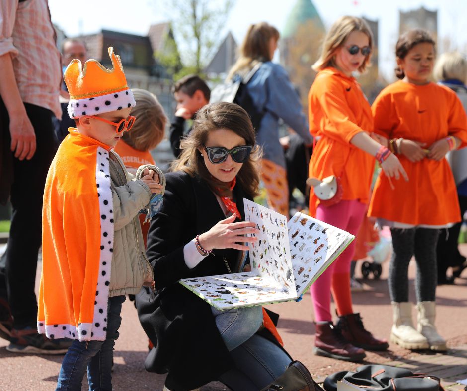 Moeder en een kind in oranje kleding kijken in een boekje met twee meiden in het oranje op de achtergrond