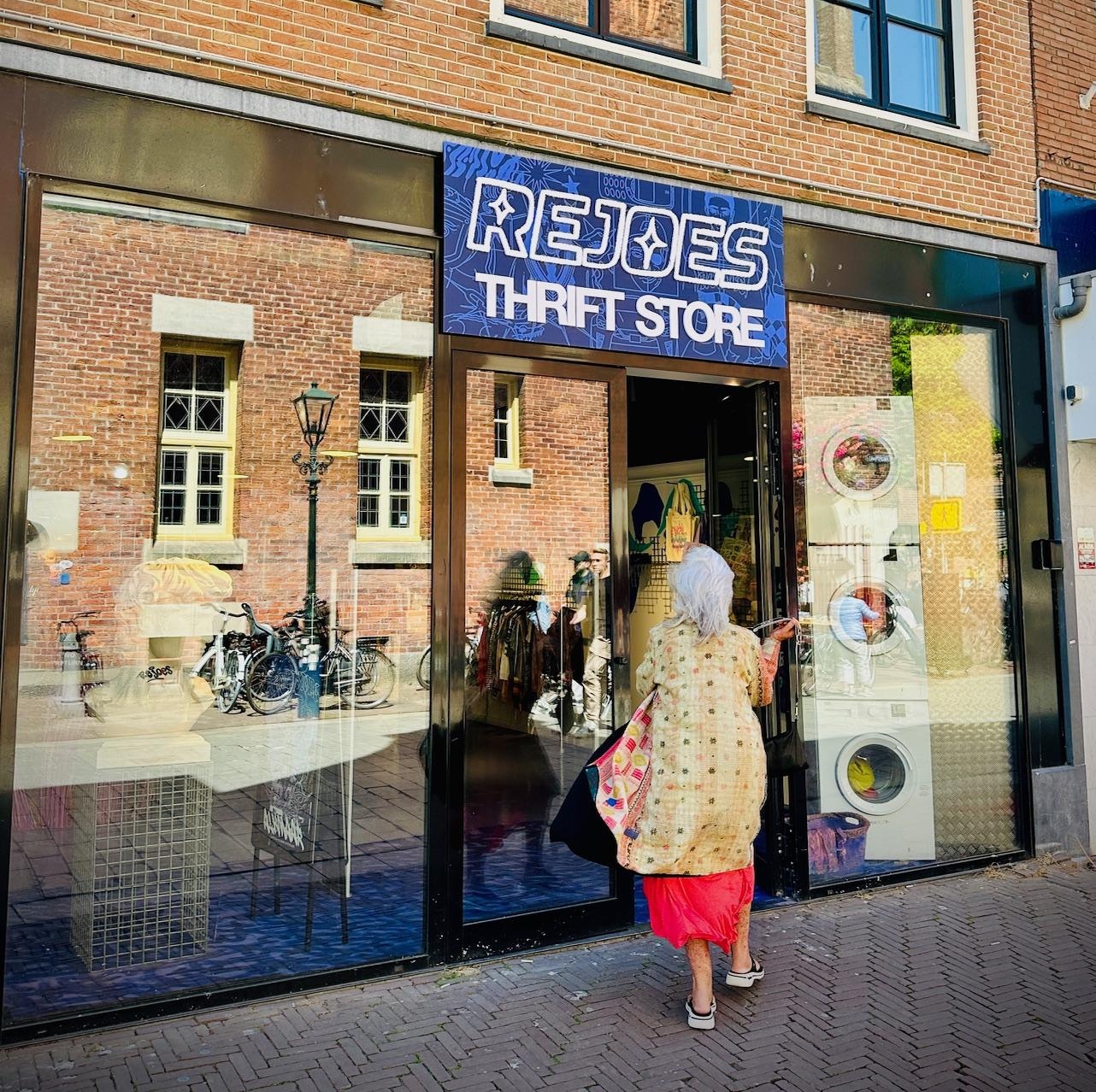 Etalage van de Rejoes Thrift Shop met het blauwe logo op de voorkant van de winkel en een vrouw die de winkel binnenkomt