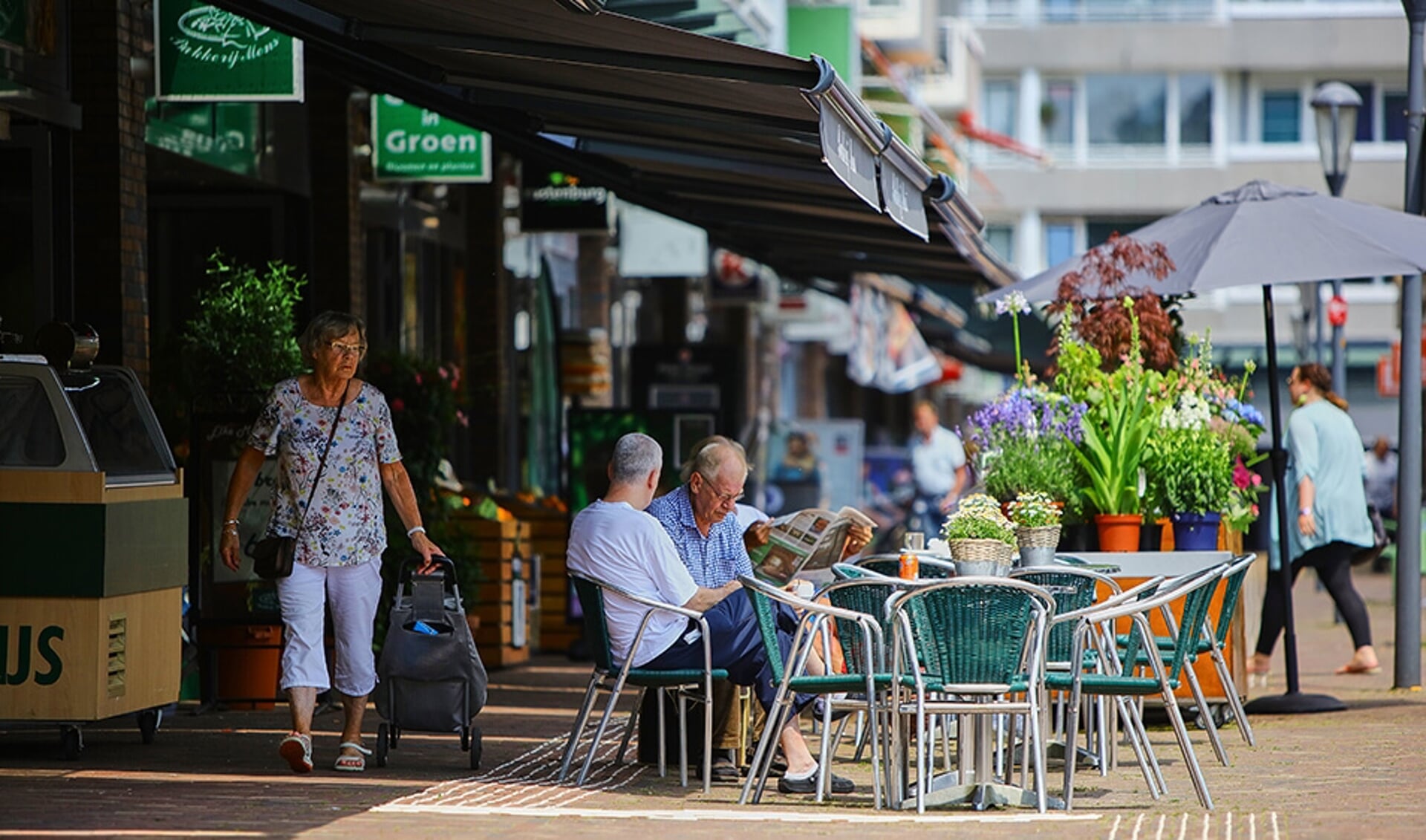 Winkelcentrum de Hoef. 2 mannen aan een tafel die zitten voor de bakker en een wandelende oudere vrouw